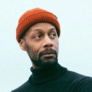 Man wearing orange knit beanie and black turtleneck, looking thoughtfully sideways against soft sky background.