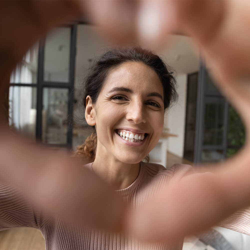 Woman smiling with healthy teeth, framing face with hands in heart shape, showcasing results of comprehensive oral care routine.